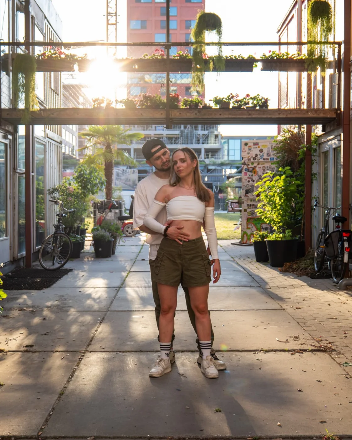 Celine and Ozi standing together in a sunlit courtyard beneath hanging plants.