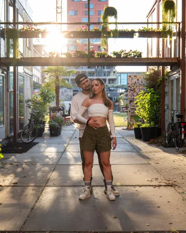 Celine and Ozi standing together in a sunlit courtyard beneath hanging plants.