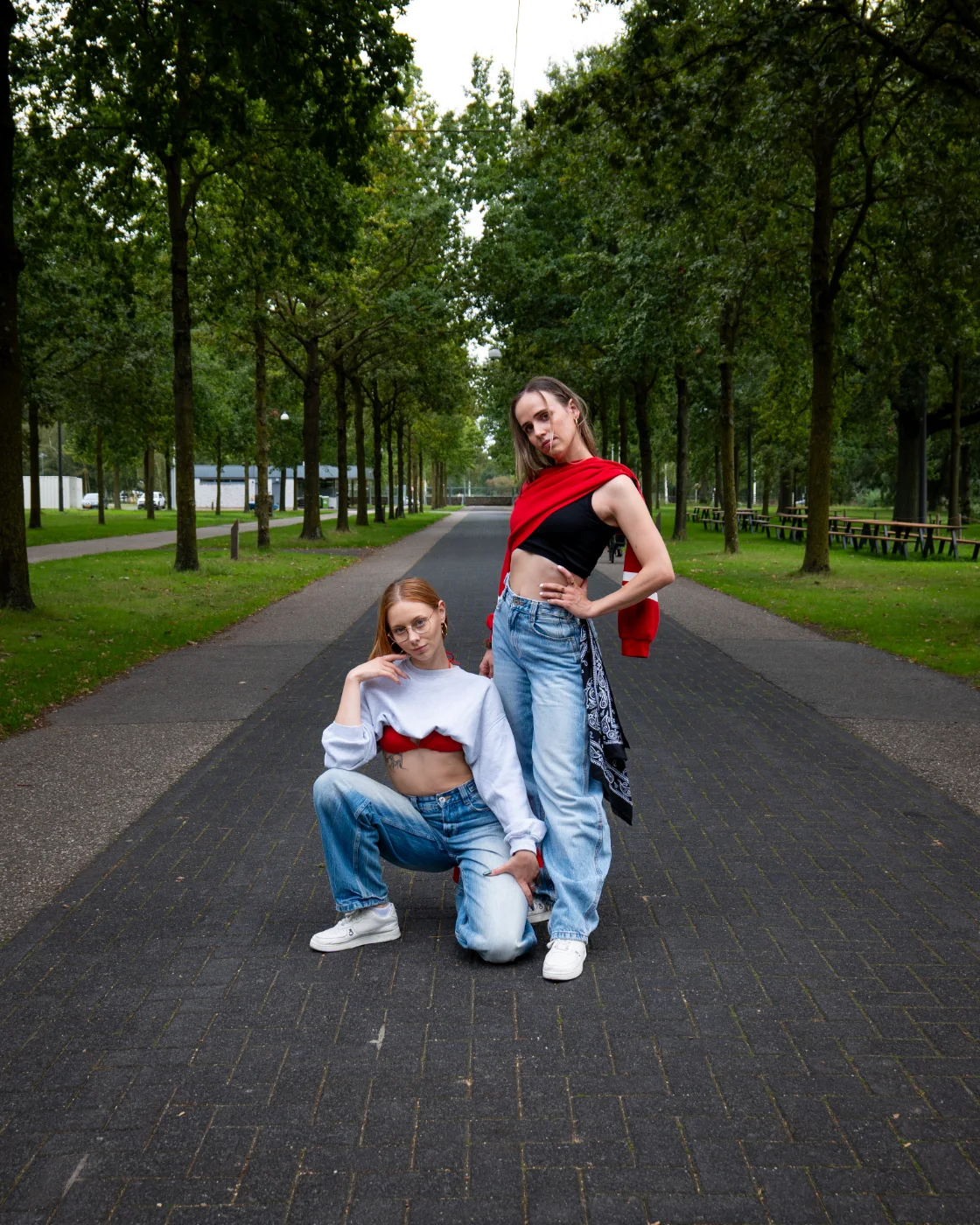 Two women posing on a tree-lined path, one crouching and one standing in blue jeans.